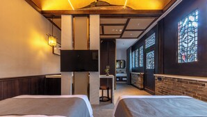 Habitación de hotel con dos camas, un televisor, una mesita con una planta y un baño visible a través de una puerta de vidrio.