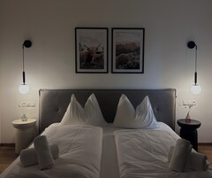 Uma cama de casal com lençóis brancos, duas mesas de cabeceira com luminárias e duas fotografias emolduradas na parede.
