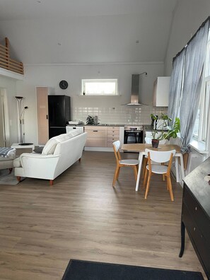 A modern living room with a sofa, dining table, and chairs. There is a kitchen area with a refrigerator, stove, and cabinets. A clock is visible on the wall. - Cozy Modern Apartment in Skellefteå - Perfect Retreat (Skelleftea)