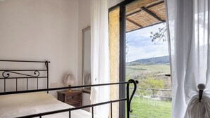 A bedroom with a metal bed frame, a bedside table with a lamp, and a view of a rural landscape through a large window. - Podere le Lune with Pool, San Gimignano (San Gimignano)