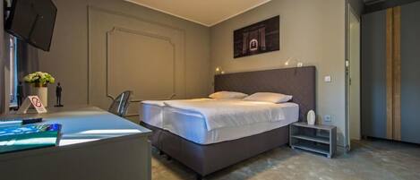 A bedroom with a double bed, a desk with a computer, a chair, and a small table with a plant.