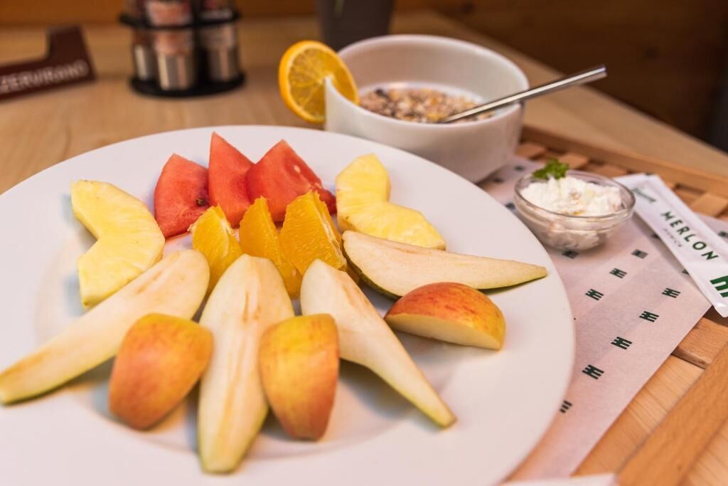 Een bord met gesneden fruit, waaronder appels, sinaasappels en watermeloen, met een kom ontbijtgranen en een klein bakje yoghurt op de achtergrond.