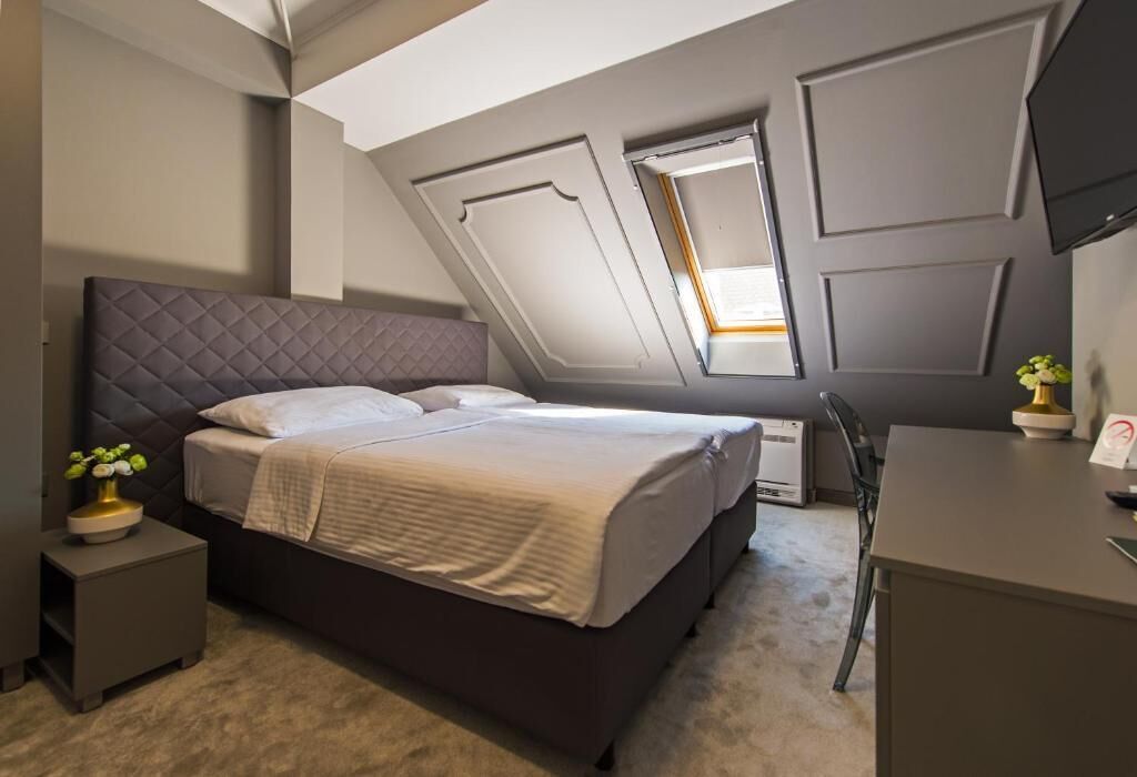 A bedroom with a bed, a desk, and a chair. There is a window with a view of the outside.