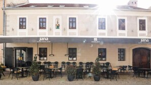 Un espace café en plein air, avec des tables et des chaises, des plantes en pot et la façade d’un bâtiment arborant le logo « jana ».