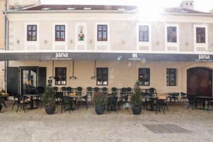 Un área de café al aire libre con mesas y sillas, plantas en macetas y la fachada de un edificio con la marca 'jana'.