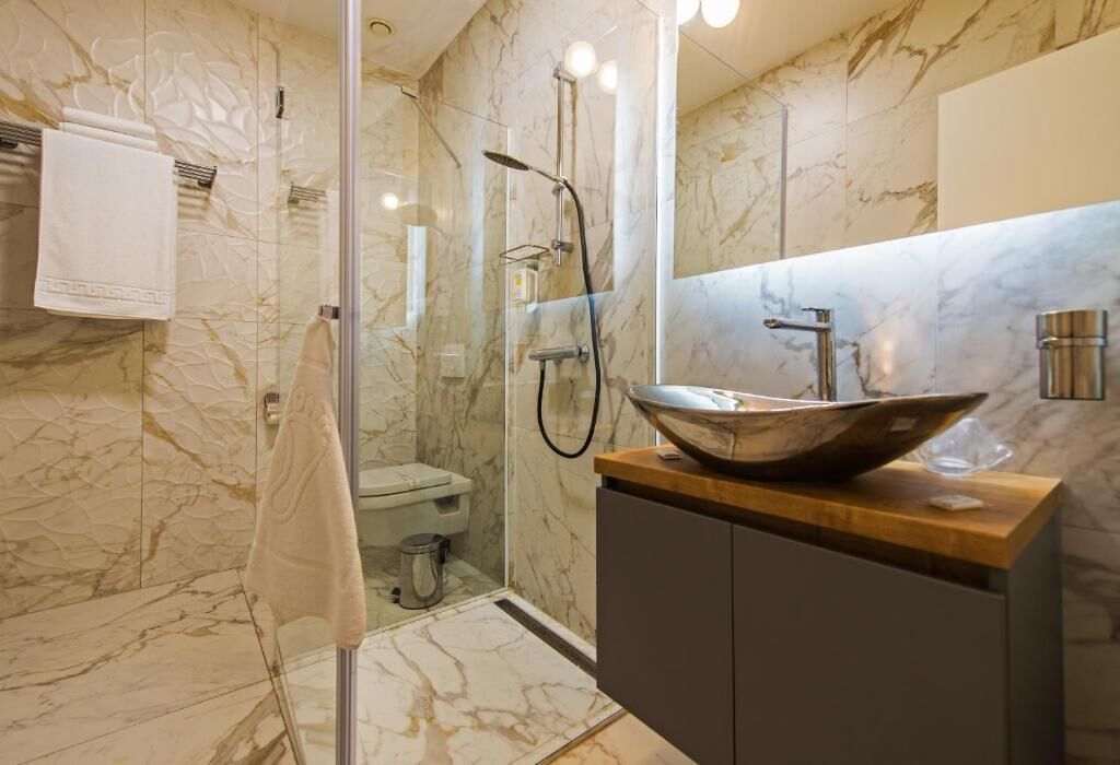 A modern bathroom with a glass-enclosed shower, a marble-tiled wall, a wooden vanity with a large vessel sink, and a toilet.