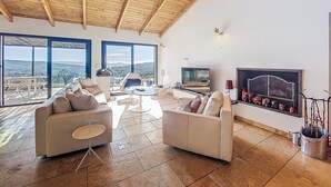 Een ruime woonkamer met een hoog houten plafond, grote ramen met uitzicht op de heuvels, een flatscreen televisie en een open haard met decoratieve objecten.