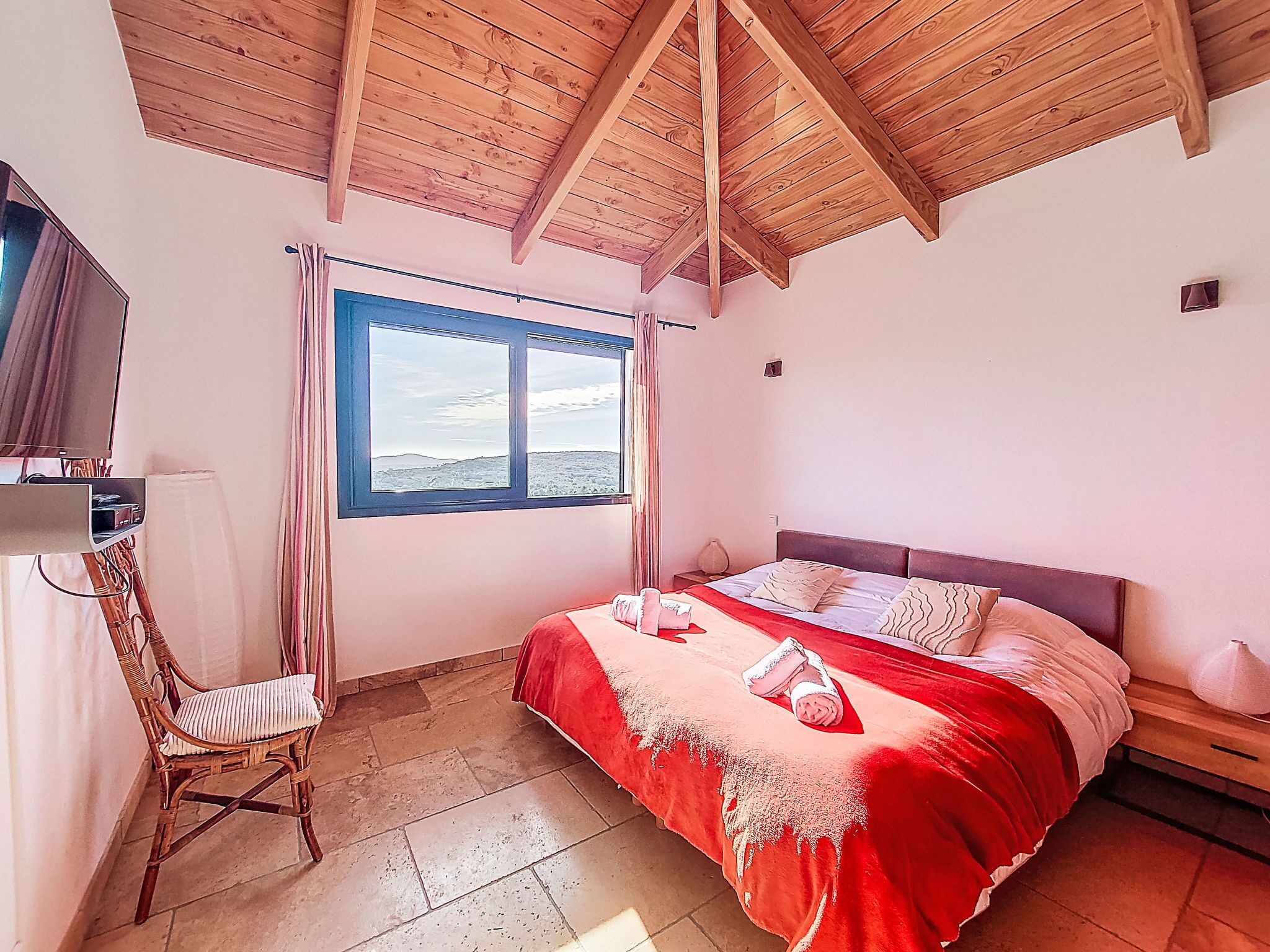 A bedroom with a wooden ceiling, a large window with a view, a bed with red bedding, a chair, and a bedside table.