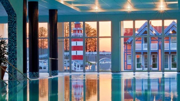 A swimming pool with clear reflections, large windows, and a view of a red and white lighthouse. - Sunday Resort Hafendorf Penthouse Suite (Rheinsberg)