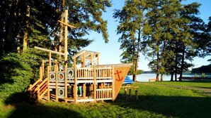 A wooden play structure with a slide and climbing elements, situated in a park with a lake and trees in the background. - Sunday Resort Hafendorf Penthouse Suite (Rheinsberg)