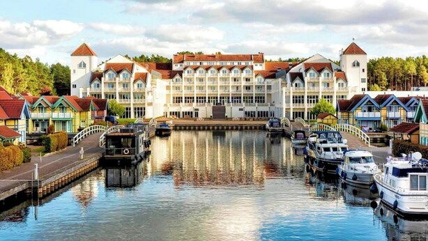 Un hôtel situé au bord d’un canal, avec un bâtiment central, des bateaux amarrés le long de la berge et des maisons colorées de part et d’autre.