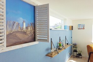 A room with a large window, a wooden shelf with decorative items, and a painting of a coastal scene. - Pristine Beachside Base in Noordwijk (Noordwijk)