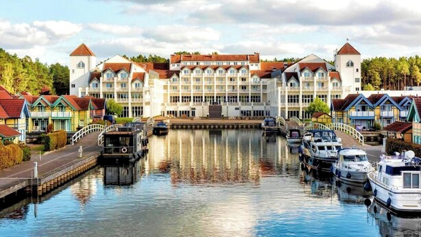 Um hotel na margem de um canal, com um edifício central, barcos atracados na água e casas coloridas de ambos os lados.