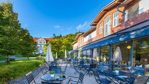 An outdoor patio with tables and chairs, a brick building with large windows, and a tree-lined path. - Sunday Resort Rügen Junior Suite Manor House (Sagard)