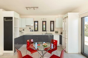 Eine moderne Küche mit einem Glastisch und roten Stühlen, eine Tafel mit dem Schriftzug 'Summer' und Blick ins Freie durch die Fenster.