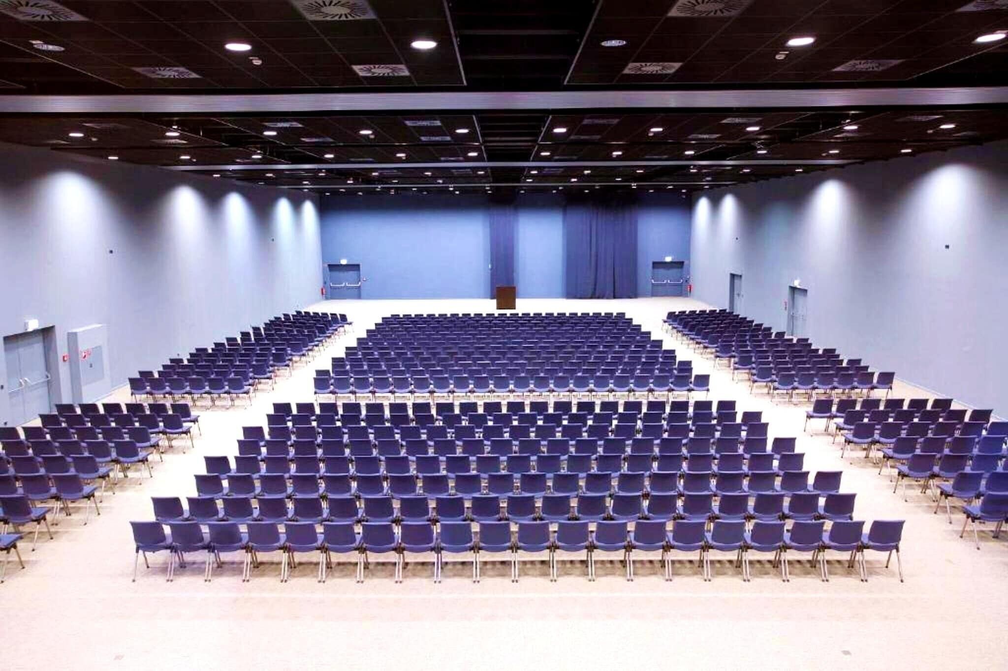 A large, empty auditorium with rows of blue chairs and a stage at the front.