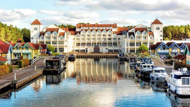 A canal-side hotel with a central building, boats docked along the water, and colorful houses on either side.