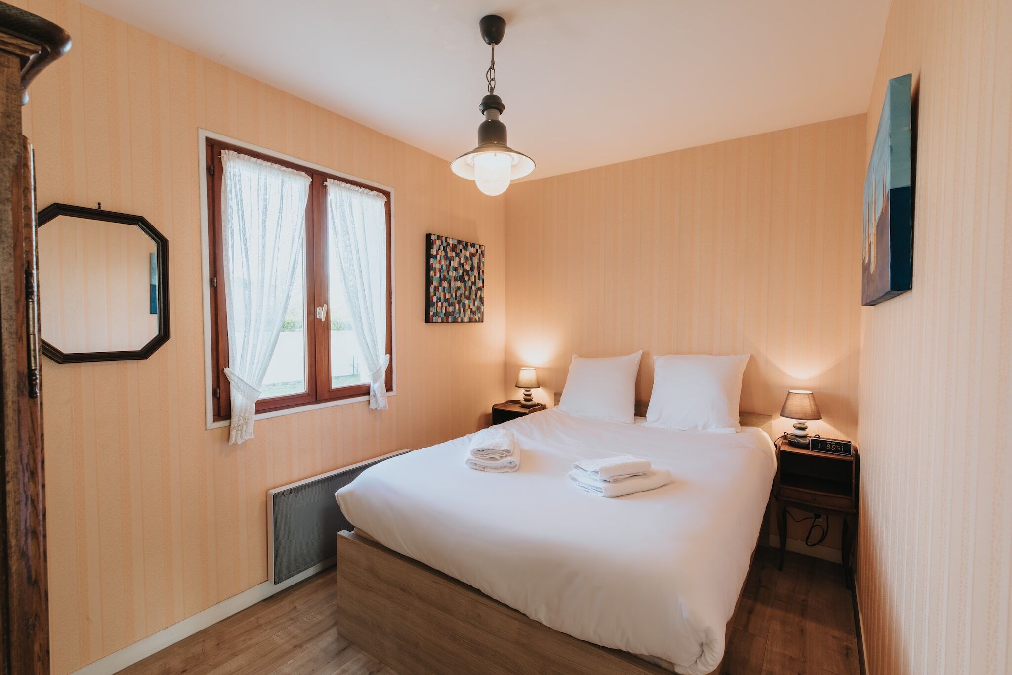 A hotel room with a bed, two bedside tables, a window with curtains, a wall-mounted artwork, and a ceiling light fixture.