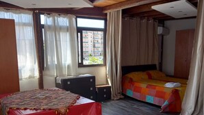 Ein Zimmer mit Holzboden, einem Bett mit farbenfroher Steppdecke, einem Tisch mit roter Tischdecke und einem Fenster mit Vorhängen.