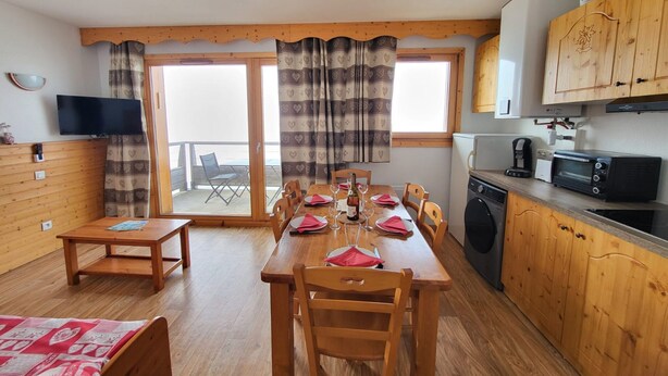 A cozy kitchen with wooden cabinets, a dining table set for four, a washing machine, and a TV mounted on the wall. - Apartment in Chamrousse Near Cable Car (Chamrousse)