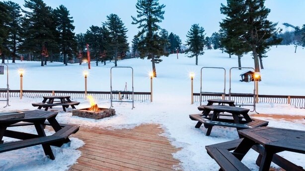 A snow-covered outdoor area with picnic tables and a fire pit, surrounded by trees and a wooden deck. - Upper Cedar Haus Retreat with Fireplace, Balcony & Mountain Views (Iron Mountain)
