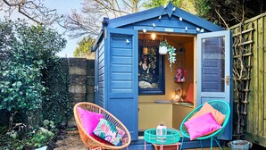 A blue shed with a door open, revealing a painting and a hanging plant. Two wicker chairs with colorful cushions are outside. - Sea Gables (Exmouth)