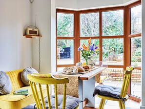 A cozy room with a yellow sofa, a wooden table with breakfast items, and a large window with a view of greenery. - Sea Gables (Exmouth)