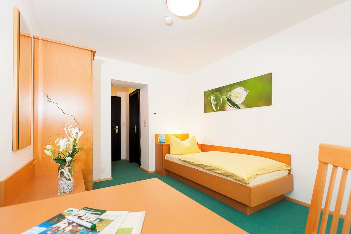 A room with a bed, a wooden chair, a desk with a vase of flowers, and a picture on the wall.