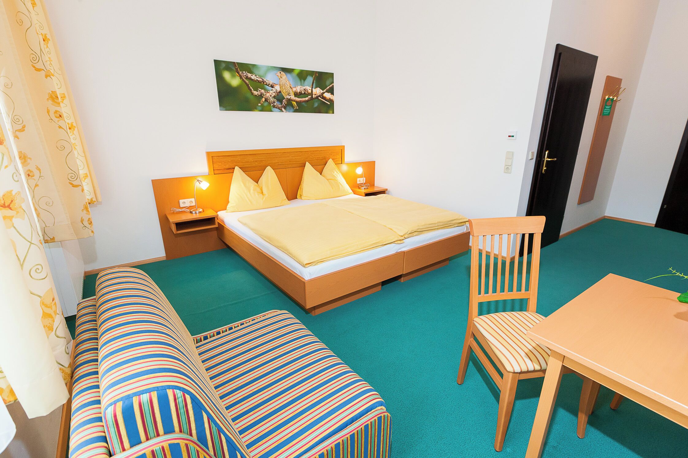 A hotel room with a bed, a wooden table, a chair, a bench, and a wall-mounted picture.