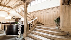 A grand staircase with wooden balusters and a reception desk with a clock and lamps. - Naturhotel Schloss Kassegg (Sankt Gallen)