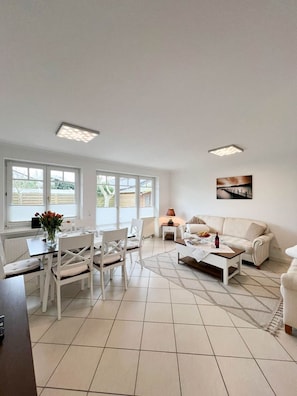 A spacious living area with a dining table, white chairs, a sofa, and a coffee table. There are large windows providing natural light and a view of the outside. - Haus Baylan - Welcome to Haus Baylan, a charming vacation home on the (Sylt)
