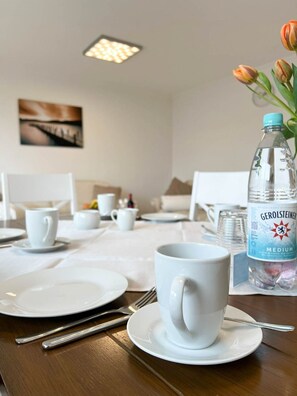 Une table de restaurant d’hôtel, dressée avec de l’assiette blanche, des tasses et des couverts, une bouteille d’eau Gerolsteiner et un vase rempli de tulipes.