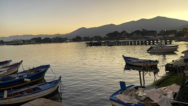 A calm river with boats docked, a pier extending into the water, and a mountain range in the background. - Laura's Little Houses: Mondello 1 (Palermo)