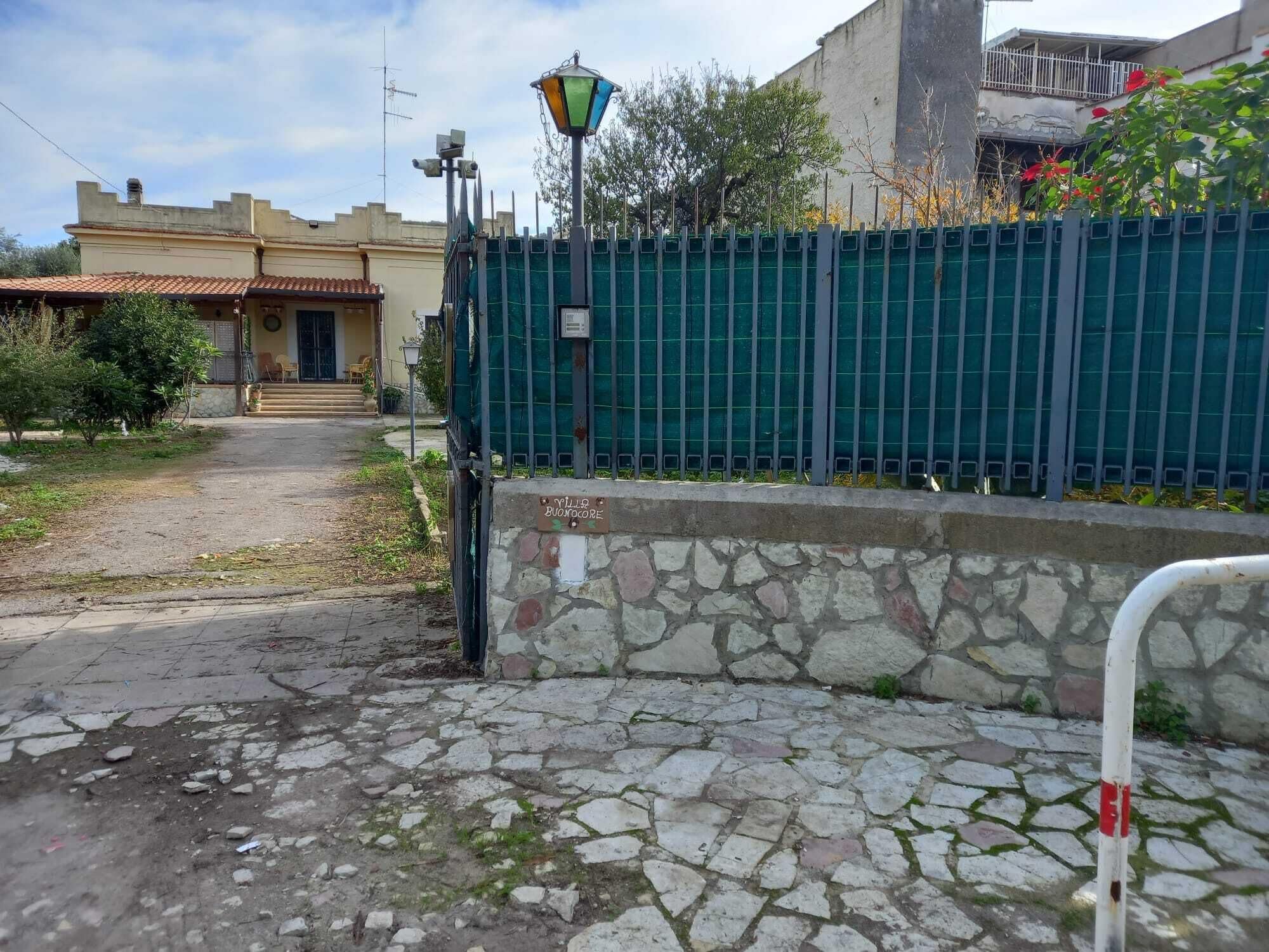 A stone-paved entrance with a metal gate, a street lamp, and a building with a tiled roof in the background.
