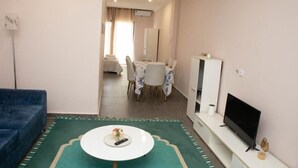 A living room with a blue sofa, a white coffee table with a remote control, a flat-screen TV, and a dining area with a table and chairs. - Nour Apatments (São Tomé)
