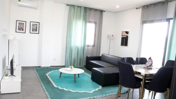 A modern living room with a black sofa, a round coffee table, and a dining area with a white table and black chairs. - Nour Apatments (São Tomé)