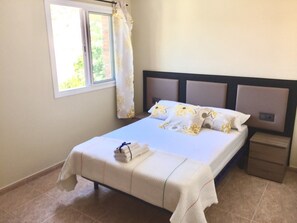 A neatly made bed with a white bedspread and matching pillows, a small bedside table, and a window with curtains. - Elegant house w/pool | Aroa by HL (barcelona)