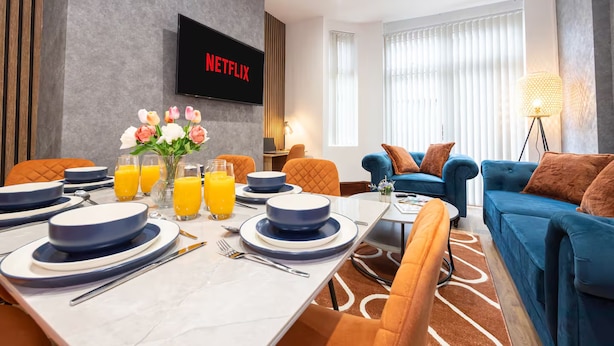 A modern hotel room with a dining area, a living area with a blue sofa, and a TV screen displaying 'NETFLIX'. - Modern & Spacious 4-Bedroom Home, Family & Contractor Friendly, Manchester (Manchester)