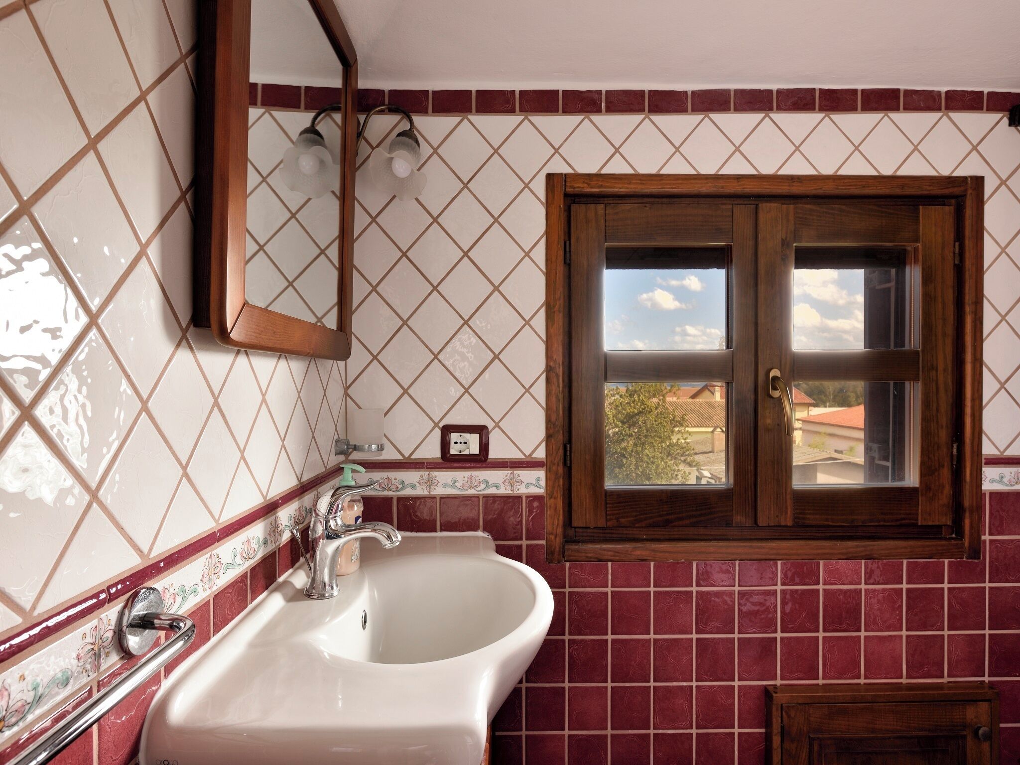Une salle de bain avec un lavabo blanc, un miroir et une fenêtre donnant sur des immeubles et des arbres.
