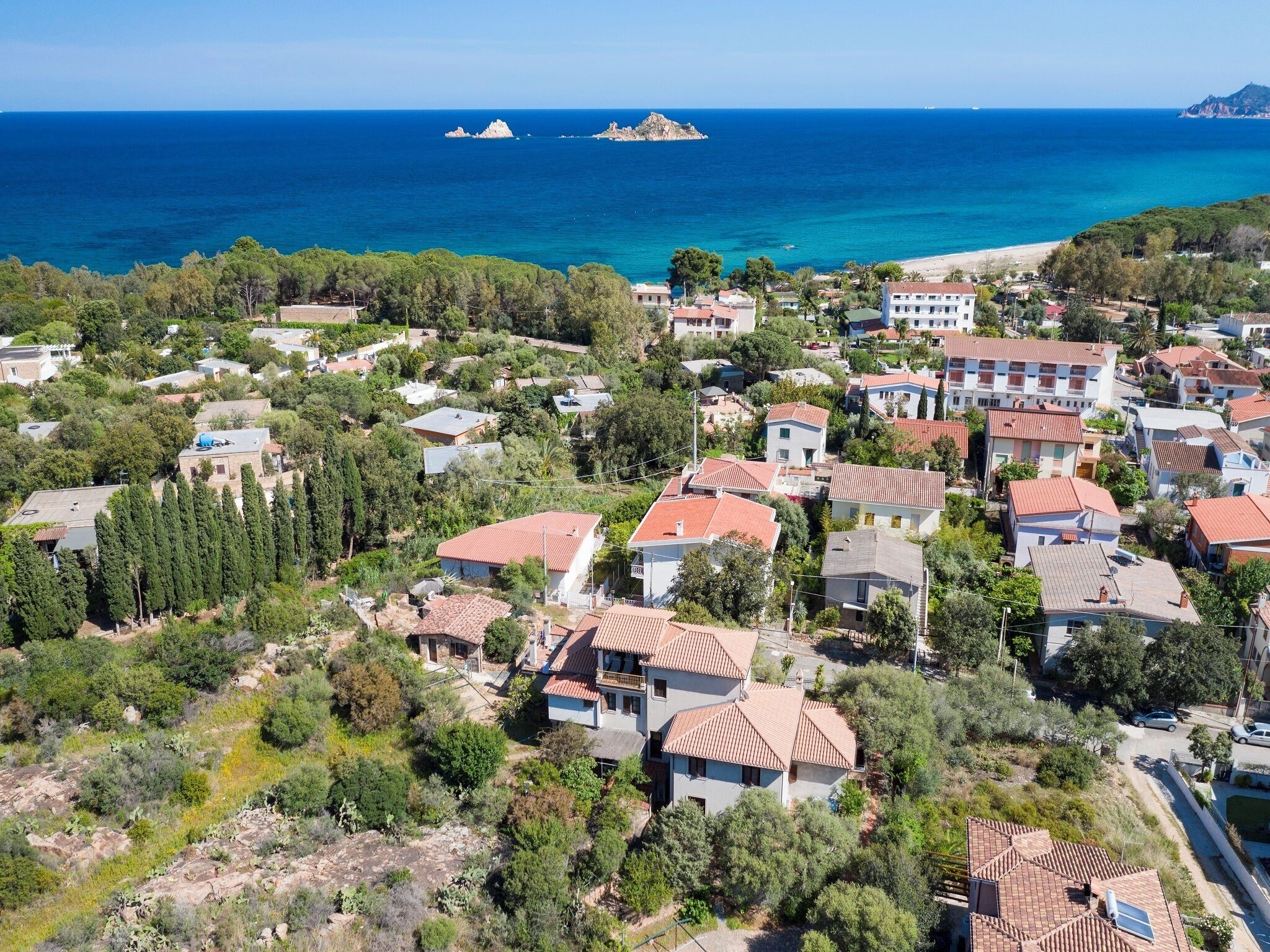 Un quartier résidentiel situé sur la côte, avec des maisons, dont certaines sont coiffées de toits en tuiles rouges, et qui offre une vue dégagée sur la mer et sur de petites îles au loin.