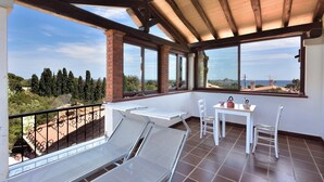 A balcony with two reclining chairs, a small table, and a chair. The balcony has large windows offering a view of the sea and surrounding landscape. - Apartment with beautiful sea and mountain views. (santa maria navarrese)