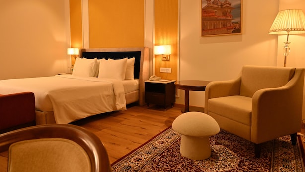 Une chambre d’hôtel avec deux lits, un coin salon comprenant un fauteuil et un pouf, une petite table et une lampe.