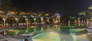 A nighttime pool area with a fountain, palm trees, and a gazebo. - Mukundra Sarovar Premiere Kota (Kota)