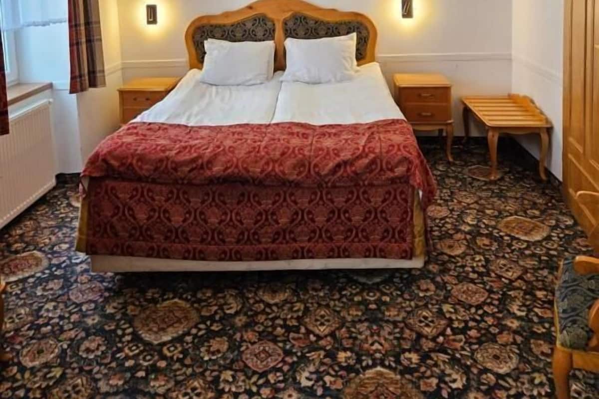 A bedroom with a large bed, two bedside tables, a wooden headboard, a painting on the wall, and a patterned carpet.