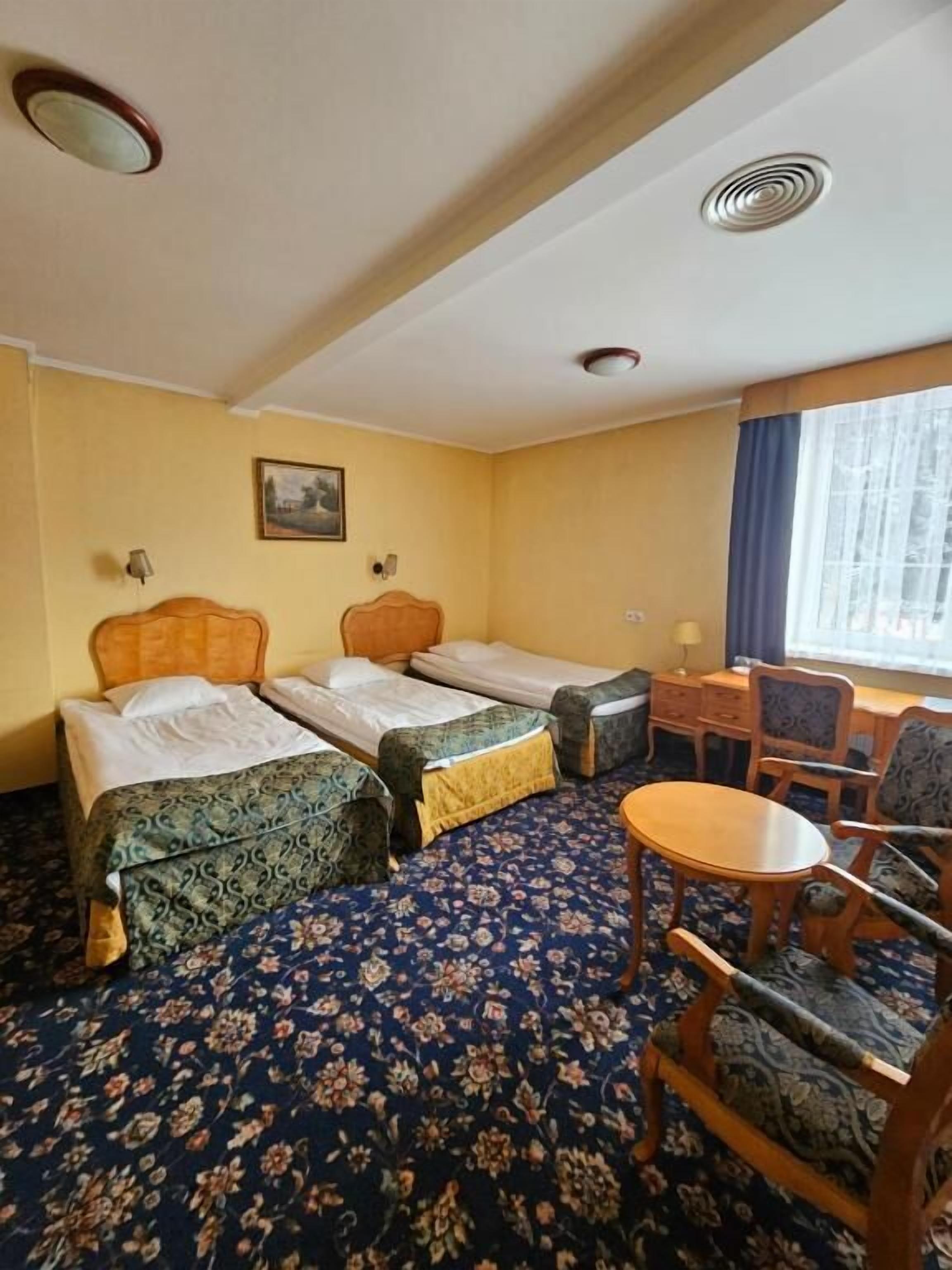 A hotel room with two single beds, a small desk with a chair, a round table, and a large window with curtains.