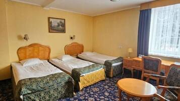 A hotel room with two single beds, a small desk with a chair, a round table, and a large window with curtains.