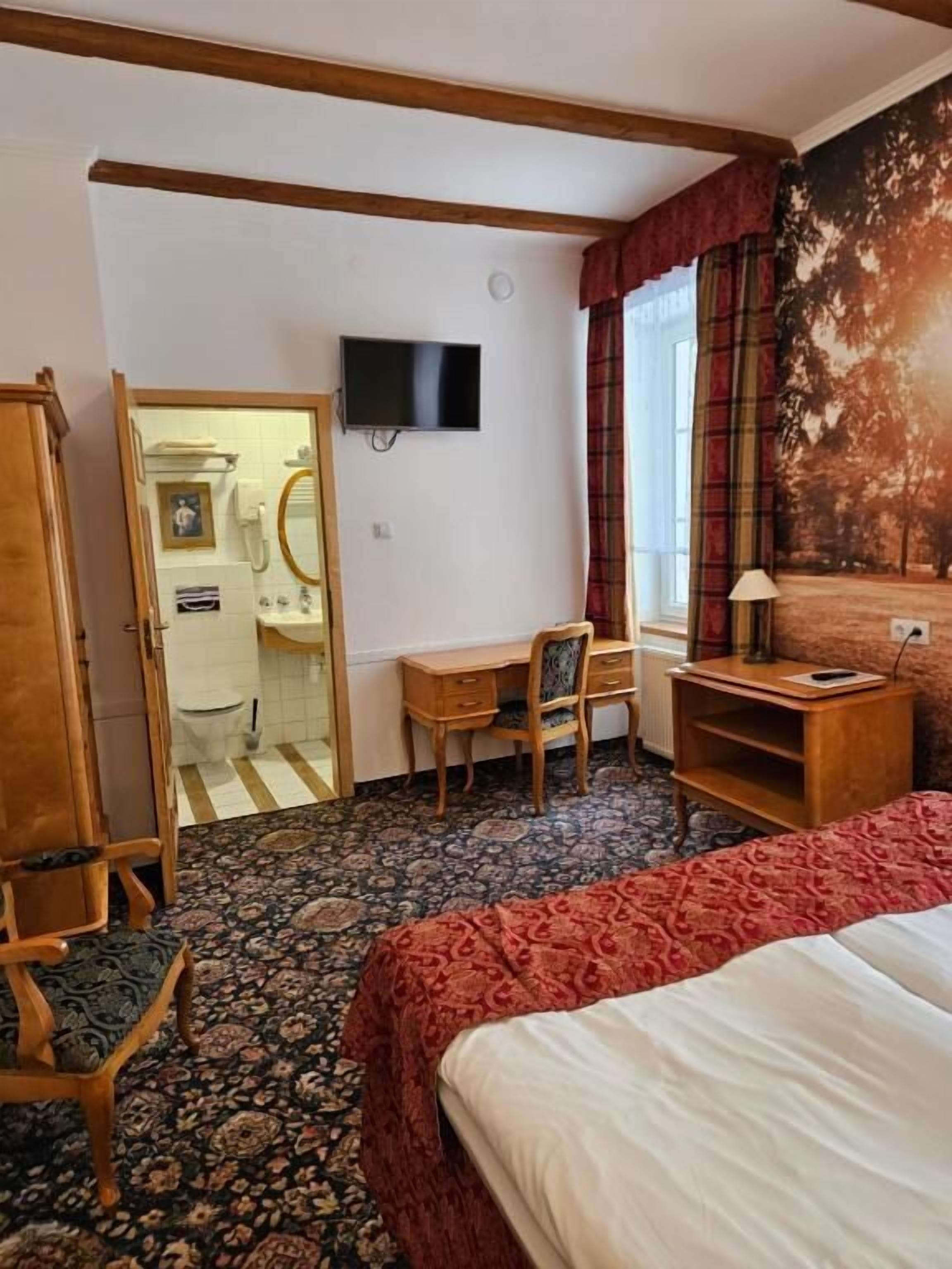 A hotel room with a bed, a desk with a chair, a television, a window with curtains, and a door leading to a bathroom.