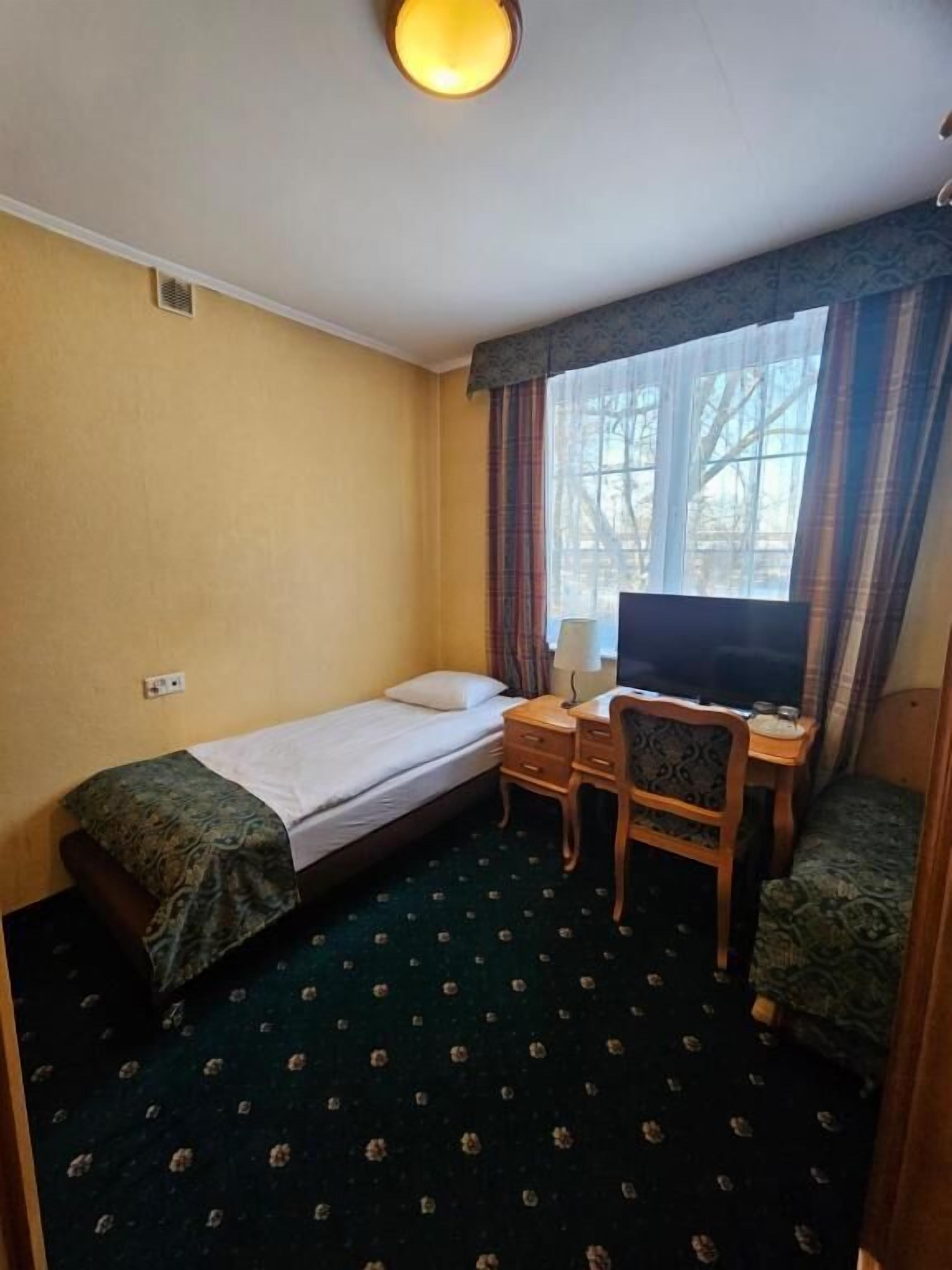 A small hotel room with a single bed, a desk with a chair, a television, and a window with curtains.