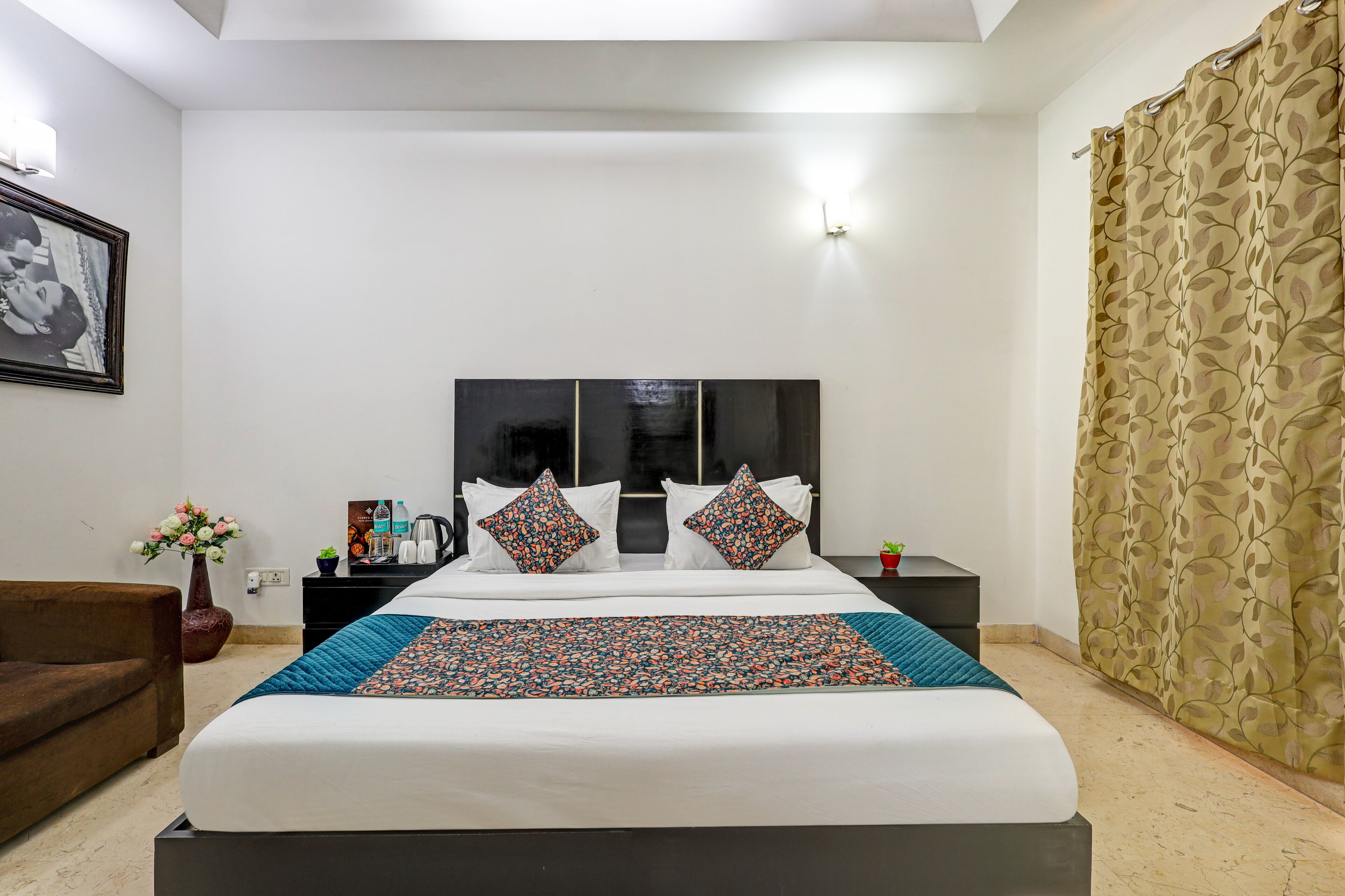 A hotel room with a large bed, a nightstand with a lamp, a chair, a framed picture on the wall, and a window with curtains.