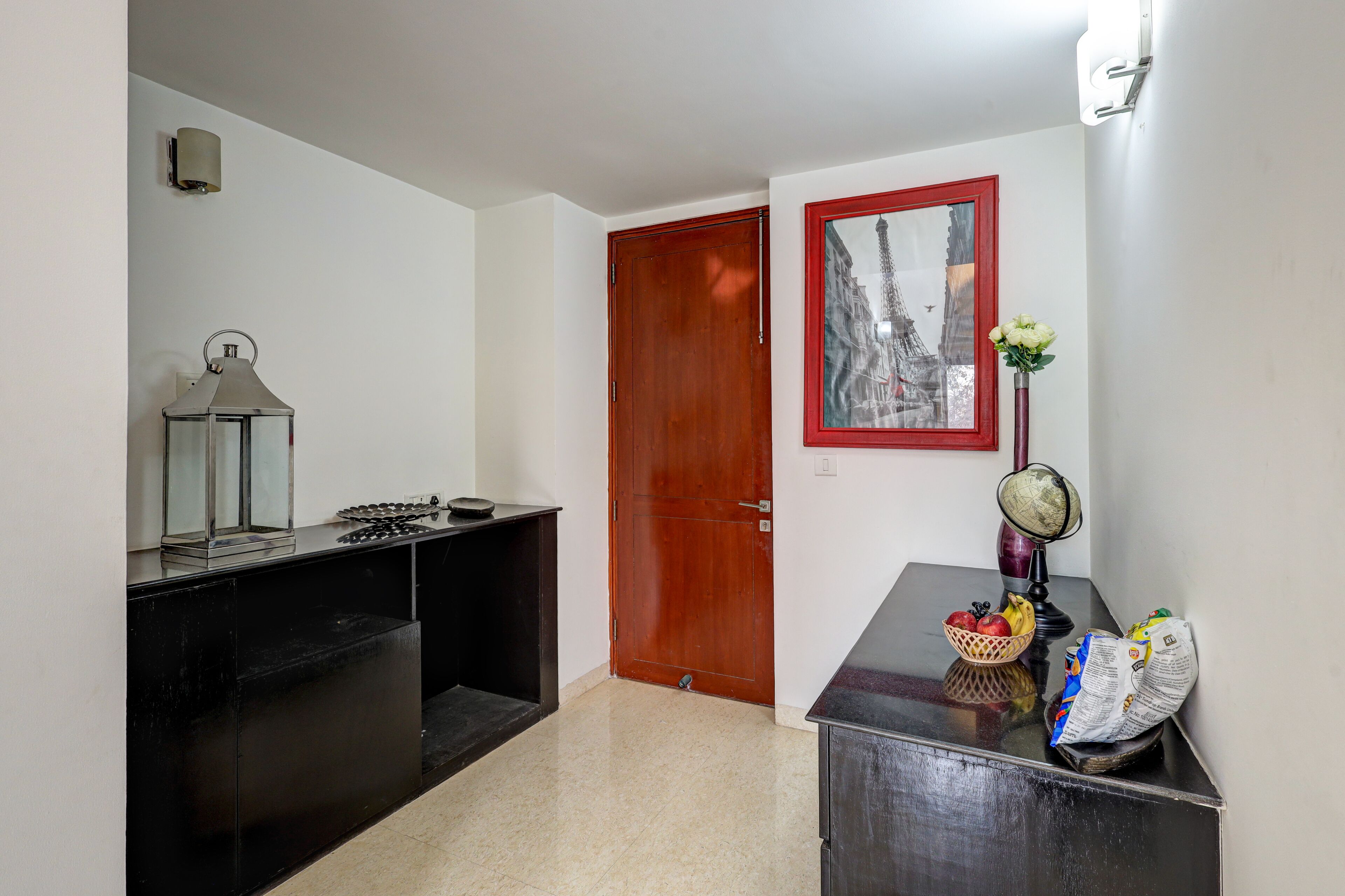 Un pasillo con una puerta de madera, un cuadro enmarcado, una mesita con un jarrón y una cesta de fruta.